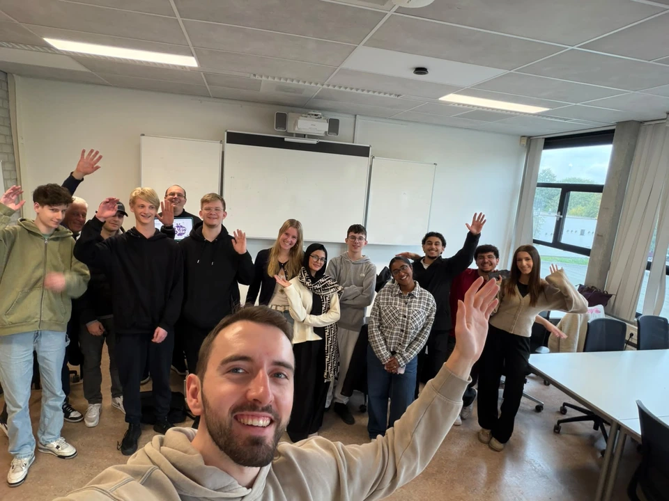 A group photograph in a classroom during the mboRijnland game jam, with several participants waving at the camera.